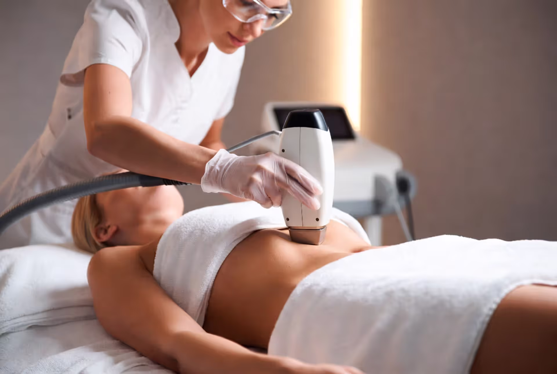 Professional Brazilian laser hair removal procedure in a modern clinic. Woman lying on treatment bed covered with white towels, technician performing the treatment
